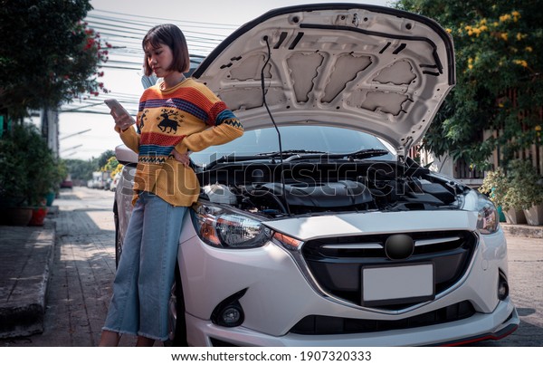 A
woman standing next to a car with a bonnet opened and using a cell
phone to contact a mechanic to fix the damaged
car.