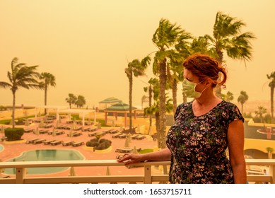 A woman is standing on a balcony in the hotel and has a face mask to protect her from the Calima sandstorm, which brings a lot of fine dust with her. Concept: health and travel