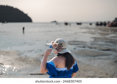 A woman standing on the beach looking out at the sea She wore a blue off-the-shoulder dress. and a wide-brimmed hat decorated with colorful flowers. - Powered by Shutterstock