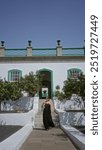 Woman standing on elegant terrace steps in front of whitewashed hotel with green accents under clear blue sky outdoors