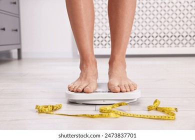 Woman standing on floor scale and measuring tape at home, closeup. Weight control