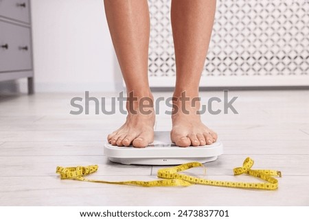 Woman standing on floor scale and measuring tape at home, closeup. Weight control