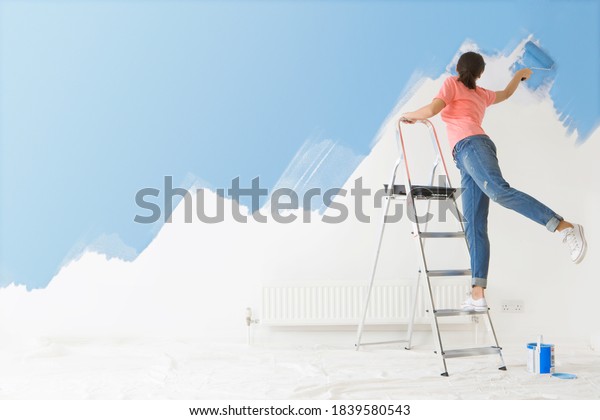 Woman standing on a ladder is applying blue\
paint to the wall in her house using a paint-roller with her back\
to the camera.\
