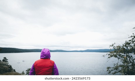 A woman standing on the rocky shore of a lake, gazing out at the water on a cloudy day, wearing a purple-red jacket, appearing contemplative, with mountains in the background and cloudy sky. - Powered by Shutterstock