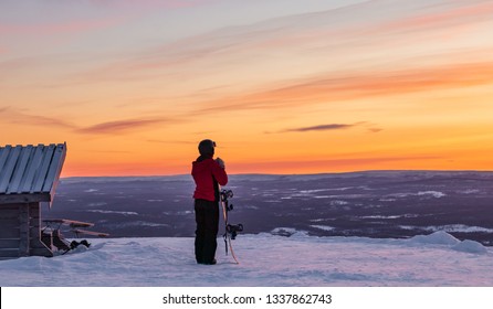 Woman standing on top of Idre Fjall, Sweden with her snowboard watching a dramatic sunset over the valley below with a frozen forest in winter. 