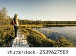 Woman standing on a wooden boardwalk, overlooking a calm lake surrounded by dense greenery as the evening sun casts a warm glow on the landscape in Idre Dalarna Sweden