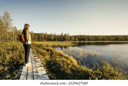 Woman standing on a wooden boardwalk, overlooking a calm lake surrounded by dense greenery as the evening sun casts a warm glow on the landscape in Idre Dalarna Sweden - Powered by Shutterstock