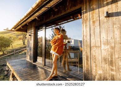 Woman standing on a wooden deck in an orange sweater, enjoying the golden morning sunlight. The reflections in the glass capture a serene and tranquil atmosphere