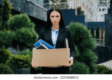 A woman standing outside, holding a box, looking lost and jobless. An unemployed woman with a lost job, holding a box of her belongings. Weight of Loss Woman Carrying Box After Being Fired