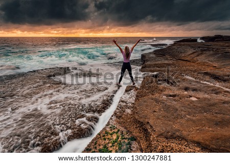 A woman standing over a rock crevice arms outstretched with excitement and zeal as powerful waves wash in over the rocks and into the chasm.