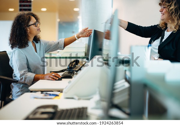 Woman standing at reception\
desk giving her card to the receptionist. Woman visiting\
municipality office being assisted by the administrator at front\
desk.