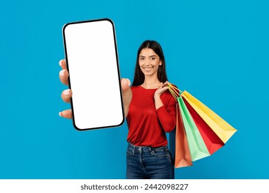 Woman standing with shopping bags in one hand and blank smartphone in the other, smiling young female multitasking while making purchases, recommending new app, mockup