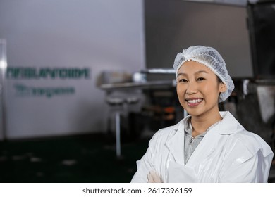 Woman standing, smiling with crossed arms in food processing plant, lab coat, hair net, copy space. Industrial, manufacturing, stainlesssteel, machinery, quality control, inspection, hygiene