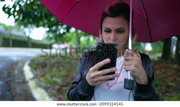 Woman standing in street holding\
umbrella looking at smartphone waiting for car\
transportation