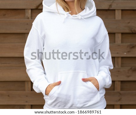 Woman is standing in white hoodie with hands in pockets in front of camera. She is posing for clothing mockup. On her hoodie is empty space for design or incription.  Sweatshirt hood template