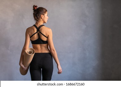 Woman standing at work out or yoga class with rolled fitness mat in her hads turned her back to the camera. Copy space on dark background