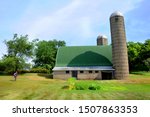 Woman stands besides a green roofed dairy barn with two tall silos.  Compared to young woman