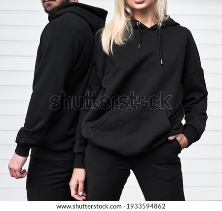Woman stands in black no logo hoodie with bearded man behind her. Clothes branding mockup. Design template for casual sportswear