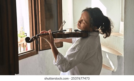 A woman stands by an open window passionately playing the violin, embracing the music's emotion. Her eyes are closed, reflecting her deep connection with the melody and natural lighting. - Powered by Shutterstock