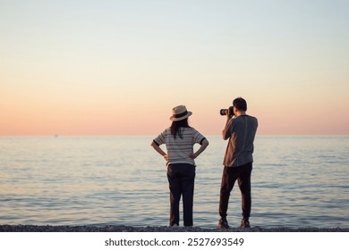 A woman stands by the sea at sunset, while a photographer captures the moment - Powered by Shutterstock