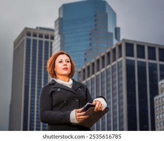 A woman stands confidently in front of towering skyscrapers, holding a tablet in her hands. The overcast sky and modern architecture create a professional and urban atmosphere.
 - Powered by Shutterstock