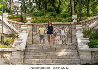 A woman stands confidently on stone stairs with two children, surrounded by lush greenery and colorful flowers in a serene garden setting - Powered by Shutterstock