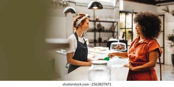 Woman stands at the counter of a coffee shop, using her credit card to make a payment with the help of a friendly barista. Happy waitress and customer doing a cashless transaction. - Powered by Shutterstock