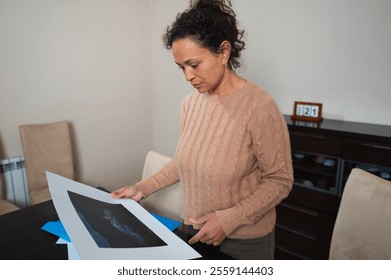 A woman stands in a cozy room, thoughtfully examining a piece of artwork. Her focus emphasizes the significance and emotion behind the art. - Powered by Shutterstock