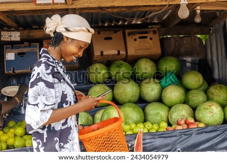 A woman stands in front of a variety of fresh fruits displayed on a market stand. She looks at mobile phone, possibly considering a purchase.