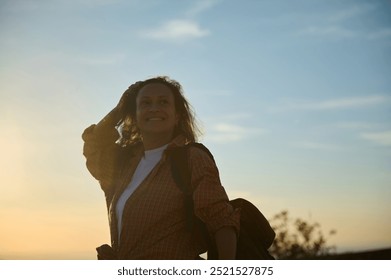 A woman stands happily against a calming sunset backdrop, with a confident smile, dressed in a flannel shirt and backpack, embodying the joy of outdoor adventure. - Powered by Shutterstock