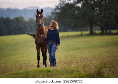 A woman stands with her horse in a meadow and looks up at the horse, smiling.
