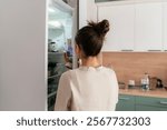 A woman stands in her kitchen opening the fridge to find something to eat during mealtime, surrounded by food items