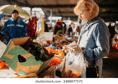 A woman stands at a market stall, inspecting a box of leafy greens while holding a plastic bag and other items. - Powered by Shutterstock