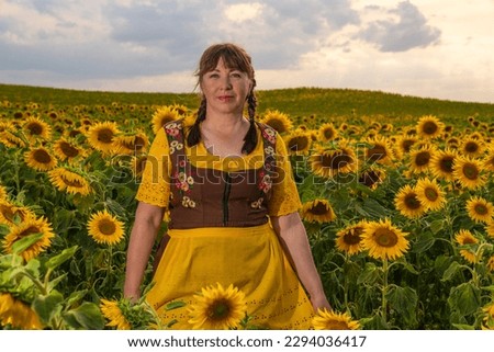 A woman stands in the middle of a field with many yellow sunflowers. Her hair is braided, dressed in German national clothes. Sunny, summer day