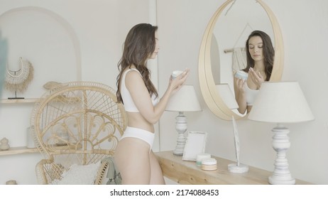 Woman stands at mirror and takes care of herself. Action. Sexy woman performs care in bright interior. Attractive well-groomed woman applies cream