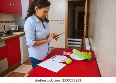 A woman stands in a modern kitchen using a tablet. She is engaged in planning a healthy meal with fresh vegetables and a cup of coffee on the counter, emphasizing healthy eating and digital lifestyle. - Powered by Shutterstock