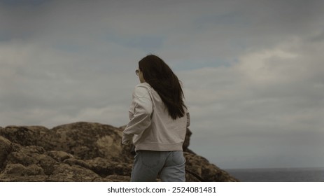 A woman stands near the rocky coastline at Castro de Baroña, Porto do Son, facing the sea. The overcast sky and rugged landscape create a mood of reflection and solitude. - Powered by Shutterstock