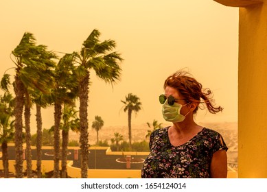 A woman stands on a balcony in the hotel and has a face mask and sunglasses to protect her from the Calima sandstorm, which brings a lot of fine dust. Concept: health and travel
