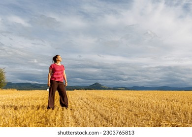 woman stands on a stubble field and stretches her face in a single ray of sunshine while rain clouds hang everywhere else