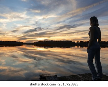 A woman stands on a wooden dock, gazing at a breathtaking sunset reflected on the calm lake waters.  - Powered by Shutterstock