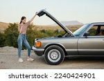 A woman stands outside a broken-down, dangerous old car with the hood open with a wrench on a road trip alone