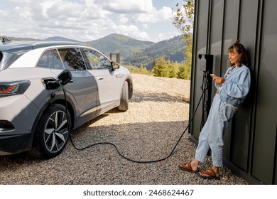 Woman stands with a smartphone and waits for electric car to be charged near a charging station at home. Concept of modern lifestyle and smart technologies