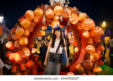 A woman stands smiling under a Halloween archway decorated with glowing jack-o'-lanterns, enjoying the vibrant holiday ambiance filled with spooky fun.
 - Powered by Shutterstock