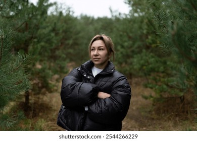 A woman stands thoughtfully with crossed arms in a lush green forest during the early hours of a cloudy day - Powered by Shutterstock