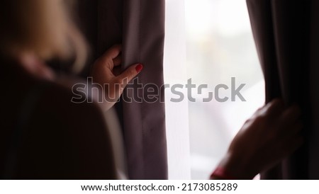 Woman stands at window and opens curtains rear view