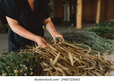 A woman stands at a wooden table in a rustic, diligently working on assembling a natural wreath from an assortment of freshly cut branches and foliage that are scattered across the work surface. - Powered by Shutterstock