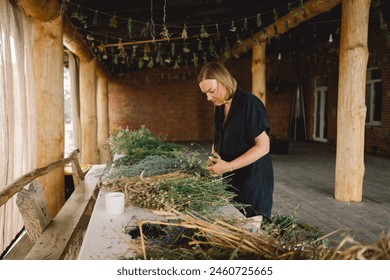 A woman stands at a wooden table in a rustic, diligently working on assembling a natural wreath from an assortment of freshly cut branches and foliage that are scattered across the work surface. - Powered by Shutterstock