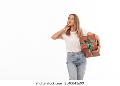 A woman stands with a wrapped present in her hands, looking thoughtfully to the side as she considers her next move. She is dressed casually, surrounded by a plain background. - Powered by Shutterstock