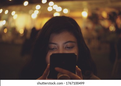 Woman Staring At Phone At Night With Blurry Crowd As Background. Woman Looking At Cellphone Screen At Night. Asian Women Staring At Smartphone At Night.