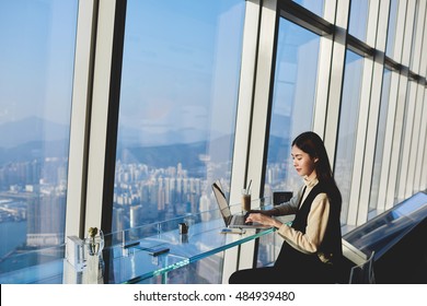 Woman Start Up Owner Of Big Prosperous Enterprise Is Working On Laptop Computer During Her Business Trip To China, While Sitting In Coffee Shop In Skyscraper Near Window With View Of Metropolitan City